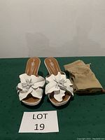 Pair of white mules with large leather hibiscus flower on top with beige fabric storage bags on a green surface, Lot 19 label visible.