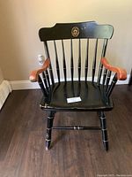 Front view of black wooden chair with gold accents and wooden armrests, showing scratches and a golden emblem on the backrest.