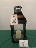 Front view of black metal electric lantern with clear glass panels and white LED candle inside, showing attached product tag.