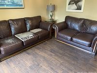 Photo showing brown leather couch and loveseat with wood plinth base, couch cushions heavily damaged with peeling leather, seating arranged in corner with lamps and framed art on walls
