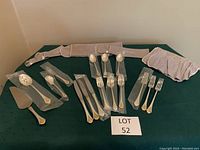 Overview of sterling silver flatware pieces arranged on a green cloth, including three place settings and extra serving utensils, some in plastic wrapping, alongside two velvet-like storage rolls.
