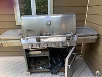 Closed view of Weber 6-burner stainless steel gas grill with thermostat on lid, six knobs on front, lower cabinet doors with one open revealing inner storage area and gas hookup connection on right side.
