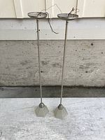 Two adjustable pendant lights with brushed metal rods and frosted glass shades standing upright on a surface with wires exposed at the top.