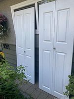 Two white bi-fold doors standing upright outside a house next to an address plaque on an exterior wall.