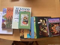 Photo showing a lineup of 8 cook books laid flat, titles include 'The Essential Vegetarian Cookbook', 'Seafood', 'Salads', 'Weight Watchers Meals in Minutes', plus two with partially hidden covers and two with unknown titles.