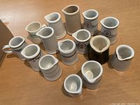 Full view of 15 small white ceramic creamers with varying decorative patterns on ceramic surfaces including floral, band and emblem details