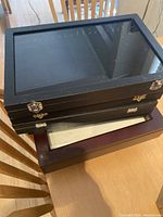 Stack of seven display cases, six black soze 14" x 9.5" x 1.75" and one larger brown 14.5" x 14.5" x 3" case under them.
