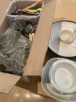Box containing clear glass sundae bowls, ceramic fish platters, cutlery, kitchen utensils, and ceramic bowls