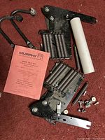 Photo of Murphy bed hardware components including metal mounting brackets with integrated coil springs, white cylindrical plastic spacer, and assorted nuts, bolts, washers. The parts rest on a red carpet.