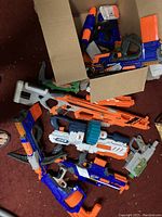 Overview of about a dozen Nerf blasters piled together showing a variety of blaster styles and colors including orange, blue, white, and gray.