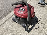 Image of the Shop Vac showing red and black body with attached suction hose and power cord on pavement.
