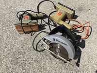 Photo showing one Black & Decker circular saw and two electric sanders on a concrete surface.