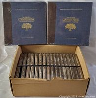 Two unopened box sets titled 'The Greatest Country Music Recordings of All Time' with a blue cover and brown spine with gold lettering, placed behind a cardboard box holding multiple cassette tapes.