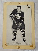 Vintage black and white photo card of hockey player Leo Boivin in Toronto Maple Leafs uniform, mounted on yellowed paper board with visible wear and tear