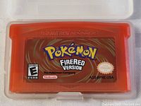 Close-up of the translucent red Pokemon Fire Red Version Game Boy Advance cartridge with label visible, placed inside a clear plastic case.