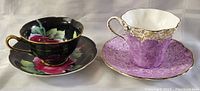 Two teacup and saucer sets, one black with floral design, one lavender with gold accents