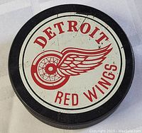 Top side of the hockey puck showing the Detroit Red Wings logo with the winged wheel and text on a white circular label on black puck.