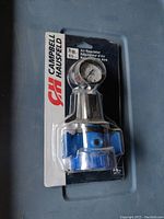 Front view of new Campbell Hausfeld air regulator sealed in original packaging. Shows blue and metal regulator body with attached pressure gauge.
