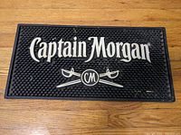 Top view of black rubber bar mat with white Captain Morgan logo and crossed swords emblem.