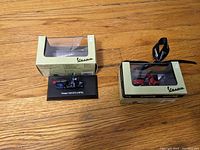Two miniature Vespa model scooters with original Vespa boxes, one model blue and one red, placed side by side on a wooden floor.