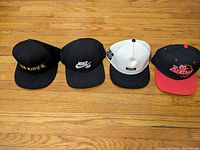 Four caps laid out side by side on wood floor: black DJ Rock, black Nike SB, white Crooks with black brim, black Adidas with red brim.