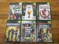 Photo showing six original Xbox game cases: Splinter Cell Double Agent, FIFA World Cup Germany 2006, Rainbow Six Critical Hour, Mercenaries Playground of Destruction, Grand Theft Auto San Andreas, Tom Clancy's Ghost Recon 2; all unused cover art visible