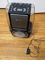 Black Singing Machine karaoke machine on wood floor, with power adapter plugged in nearby.
