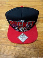 Front view of the black and red Team Rocket cap with embroidery of Team Rocket characters Jessie, Meowth, and James and 'TEAM ROCKET' text.