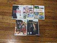 Photo shows all five Wii game cases including Scarface, Cooking Mama, Wii Play, Guilty Gear XX Core, and Goldeneye 007 laid out on wooden floor.