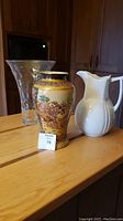 Photo of three vases on wooden table: clear glass vase, decorated Asian style ceramic vase, and white ceramic ewer.