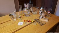 Overall lot of assorted vintage barware set on wooden table showcasing glass ice buckets, glass cocktail shaker without lid, and various small glasses.
