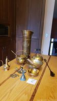Photo showing assortment of brass and copper items including Dutch coal scuttle, kettle, mulling pot with wooden pestle, and other small brass decorative items on wooden table.