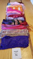 View of all 14 scarves folded and stacked on a wooden table showing vibrant colors including pinks, oranges, purples and reds.