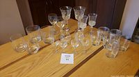 Photo shows a collection of 20 clear drinking glasses arranged on a wooden surface including stemmed glasses and short tumblers.