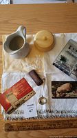 Photo of entire lot items arranged on a small embroidered yellow and cream table cover with fringe. Visible items include stoneware jug, celluloid trinket box, small silver ladle, cylindrical needle case, secret hideout book, and macabre postcard. A ruler is placed for scale.
