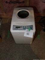 Front and top view of the white breadmaker showing viewing window and control buttons. Unit has a '90' tag attached.