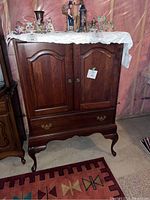 Front view of wooden cabinet with paneled double doors, curved legs, and drawer at bottom. Items on top not included.