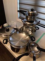 Six stainless steel pots of various sizes with matching lids and a colander insert, all with black handles, displayed on a table.