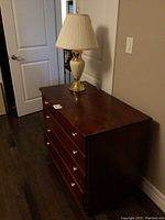Wooden 4-drawer dresser with brass-colored knobs, cream and gold lamp with pleated fabric shade on top, photographed from the side.
