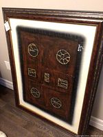 Photo of wood framed art with geometric motifs on brown background, showing the full piece leaning against a wall.