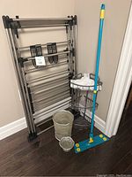 Photo showing full lot including the drying rack folded and standing against the wall, blue dry mop in front, metal corner rack with white bags, and decorative garbage cans on the floor.
