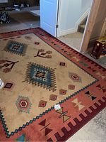 Photo showing full rug with geometric tribal patterns, cream center, rust border with multiple colored geometric motifs, placed on concrete floor near stairs.