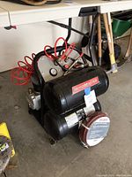 Image showing MotoMaster air compressor with dual black air tanks, attached red coil air hose, and Pro Point air hose in packaging resting in front.