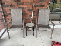 Two outdoor chairs with metal frames and floral patterned mesh seats and backs, small round side table between them, outdoor broom leaning on the wall behind.
