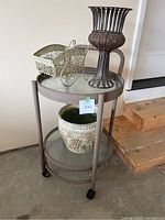 Two-tiered round metal framed serving cart with glass shelves on caster wheels holding three distinctive planters: dark brown woven metal and wicker planter, ornamental small metal planter with scrollwork, and a ceramic planter with grapevine design underneath.
