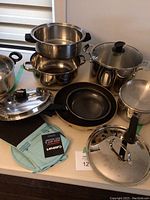 Photo showing 4 different-sized stainless steel pots with lids, 2 frying pans, a large pressure cooker, and Cuisinart oven mitts and pot holder in light blue color.