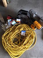 Photo showing long yellow extension cable coiled, black cord reel, cordless drill in carrying case, wiring devices, and door stops arranged on floor.