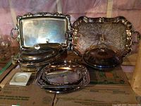 Photo showing multiple silver-plated serving trays, serving dishes with glass inserts and lids, and a pair of candle holders arranged on a cardboard surface.