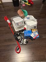 Overview of items showing Panasonic cordless phones, a digital blood pressure machine, various plastic storage containers, step stool, cleaning tools like broom, mop, dustpan, and a green plug-in wall candle with packages of light bulbs.