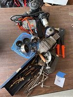Electric drill, paint stripper, drill bits, clamps, vice, chalk line, and misc. tools arranged on a wooden table with power cords visible. Tools show signs of previous use and wear.